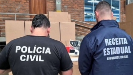 Agentes de costas, de frente para caixas de papelão. Em suas camisetas, está escrito Polícia Civil e Receita Estadual