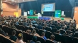 Crianças em um auditório assistem a um homem no palco. No palco, há um cartaz que diz Palestra Musicada.
