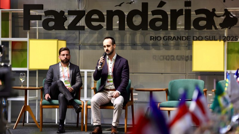 Dois homens est&atilde;o sentados em cadeiras no palco de um evento identificado pelo letreiro &ldquo;Semana Fazend&aacute;ria &ndash; Rio Grande do Sul 2025&rdquo;, vis&iacute;vel ao fundo. Um deles segura um microfone e fala ao p&uacute;blico, enquanto o outro ouve atentamente. Ambos vestem terno e crach&aacute; de identifica&ccedil;&atilde;o. &Agrave; frente do palco, h&aacute; bandeiras de diversos estados brasileiros parcialmente desfocadas. O ambiente &eacute; iluminado e apresenta um cen&aacute;rio moderno, com poltronas e pain&eacute;is coloridos ao fundo.