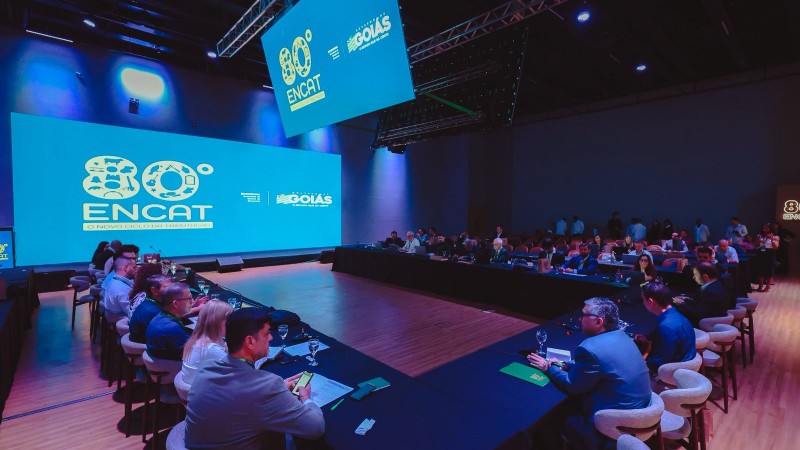 A imagem mostra um grande audit&oacute;rio moderno, iluminado com luzes em tons de azul. No centro do espa&ccedil;o, h&aacute; mesas em formato de &ldquo;U&rdquo;, onde diversas pessoas est&atilde;o sentadas, participando de uma reuni&atilde;o ou confer&ecirc;ncia. &Agrave; esquerda, ocupa boa parte da parede um tel&atilde;o gigante exibindo o texto &ldquo;80&ordm; ENCAT&rdquo; acompanhado de logotipos institucionais. No teto, h&aacute; pain&eacute;is de LED suspensos refor&ccedil;ando a identidade visual do evento. As pessoas est&atilde;o concentradas, muitas com laptops, cadernos ou tablets &agrave; frente, indicando um encontro t&eacute;cnico ou profissional. O ambiente &eacute; amplo, com piso de madeira clara e atmosfera formal.