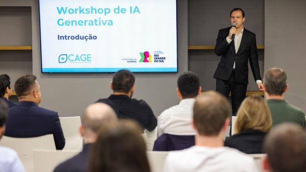 Foto de um homem em p&eacute;, vestindo terno escuro e segurando um microfone, enquanto fala para uma plateia. Ele est&aacute; posicionado &agrave; direita da imagem. &Agrave; esquerda, h&aacute; uma tela grande exibindo o t&iacute;tulo &ldquo;Workshop de IA Generativa &ndash; Introdu&ccedil;&atilde;o&rdquo;, acompanhada dos logotipos da Secretaria da Fazenda, do Governo do Rio Grande do Sul e da CAGE. A plateia est&aacute; sentada de costas para a c&acirc;mera, composta por homens e mulheres que observam a apresenta&ccedil;&atilde;o em um ambiente interno de evento.