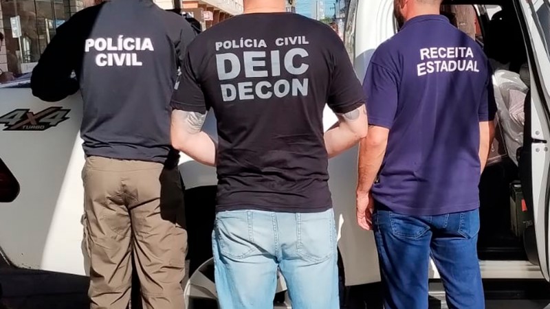 Tr&ecirc;s homens aparecem de costas, em p&eacute;, ao lado de um ve&iacute;culo branco estacionado na rua. Eles vestem camisetas com identifica&ccedil;&atilde;o institucional: &agrave; esquerda, &ldquo;Pol&iacute;cia Civil&rdquo;; ao centro, &ldquo;Pol&iacute;cia Civil &ndash; DEIC DECON&rdquo;; e &agrave; direita, &ldquo;Receita Estadual&rdquo;. O homem do centro usa camiseta preta e cal&ccedil;a jeans clara; o da esquerda usa camiseta escura e cal&ccedil;a bege; e o da direita usa camiseta azul escura e cal&ccedil;a jeans. Ao fundo, &eacute; poss&iacute;vel ver parte de pr&eacute;dios e do ambiente urbano, indicando que a cena ocorre em via p&uacute;blica durante o dia.