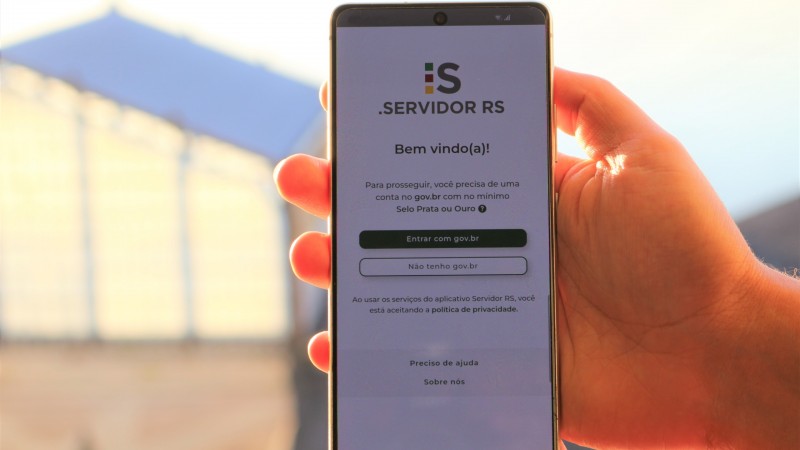 Uma m&atilde;o segura um smartphone na posi&ccedil;&atilde;o vertical, ao ar livre, com a luz do sol iluminando o fundo. Na tela do celular aparece a p&aacute;gina inicial do aplicativo Servidor RS. No topo est&aacute; o logotipo &ldquo;i:s Servidor RS&rdquo; e a mensagem &ldquo;Bem-vindo(a)!&rdquo;. Abaixo, l&ecirc;-se que, para prosseguir, &eacute; necess&aacute;rio ter uma conta no gov.br com n&iacute;vel Selo Prata ou Ouro. H&aacute; dois bot&otilde;es: &ldquo;Entrar com gov.br&rdquo; (em destaque) e &ldquo;N&atilde;o tenho gov.br&rdquo;. Na parte inferior, h&aacute; um aviso sobre pol&iacute;tica de privacidade e um link &ldquo;Preciso de ajuda&rdquo;. O fundo da imagem &eacute; desfocado, sugerindo um ambiente externo com uma estrutura clara ao longe.