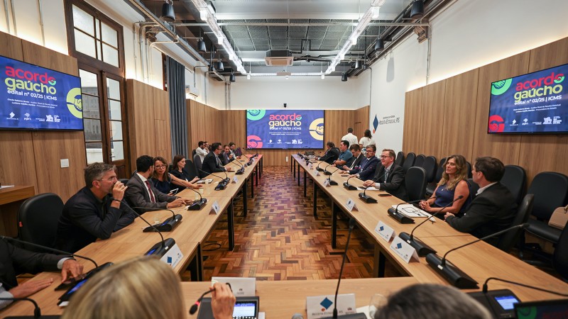 Sala de reuni&otilde;es ampla, com mesas retangulares dispostas em formato de U e microfones individuais. Em torno das mesas, h&aacute; v&aacute;rias pessoas sentadas, em sua maioria vestindo trajes formais, participando de uma reuni&atilde;o. Ao fundo, uma tela exibe o logotipo e o texto &ldquo;Acordo Ga&uacute;cho&rdquo;. O ambiente tem piso de madeira, paredes com pain&eacute;is de madeira clara e ilumina&ccedil;&atilde;o no teto, transmitindo um contexto institucional e formal.