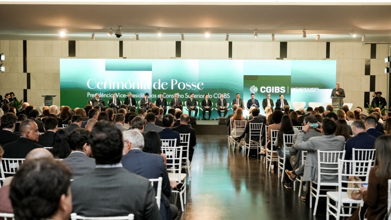 Ambiente interno amplo e bem iluminado, com um audit&oacute;rio montado para uma cerim&ocirc;nia formal. No fundo, h&aacute; um palco com um painel grande em tons de verde e branco, onde se l&ecirc; &ldquo;Cerim&ocirc;nia de Posse&rdquo; e a sigla &ldquo;CGIBS&rdquo;. Sobre o palco, cerca de dez pessoas est&atilde;o sentadas em cadeiras, dispostas em linha, vestindo trajes formais. &Agrave; direita, uma pessoa est&aacute; em p&eacute;, em um p&uacute;lpito, aparentemente discursando.

Na plateia, dezenas de pessoas est&atilde;o sentadas em cadeiras brancas organizadas em fileiras, todas voltadas para o palco. A maioria dos participantes veste roupas sociais, como ternos e blazers. A imagem &eacute; captada a partir do fundo da sala, mostrando as costas de parte do p&uacute;blico e oferecendo uma vis&atilde;o geral do evento, que transmite um clima institucional e solene.