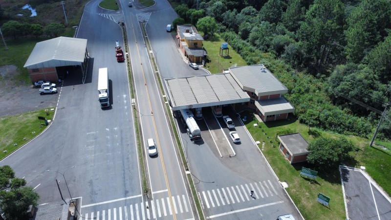 Imagem a&eacute;rea de uma pra&ccedil;a de ped&aacute;gio localizada em uma rodovia de pista dupla. No centro, um canteiro divide os sentidos da via. &Agrave; direita, h&aacute; cabines de ped&aacute;gio cobertas por uma estrutura com telhado, por onde passam caminh&otilde;es e autom&oacute;veis. &Agrave; esquerda, uma ampla &aacute;rea pavimentada serve de acesso para ve&iacute;culos de grande porte, com um caminh&atilde;o estacionado e outro em circula&ccedil;&atilde;o. Tamb&eacute;m &eacute; poss&iacute;vel ver faixas de pedestres pintadas no asfalto, pequenas edifica&ccedil;&otilde;es de apoio e uma extensa &aacute;rea de vegeta&ccedil;&atilde;o e &aacute;rvores ao redor da rodovia.